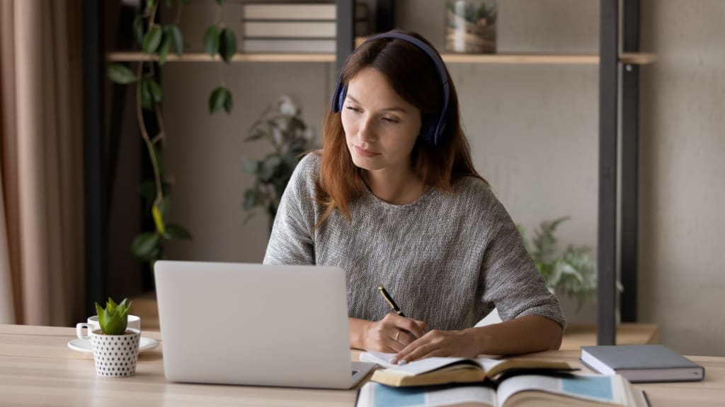 A woman wearing headphones writes in a notebook while consulting a laptop, working on a mental health website design. She is seated at a desk adorned with books, a plant, and a mug. Shelves filled with plants and books create a serene backdrop.